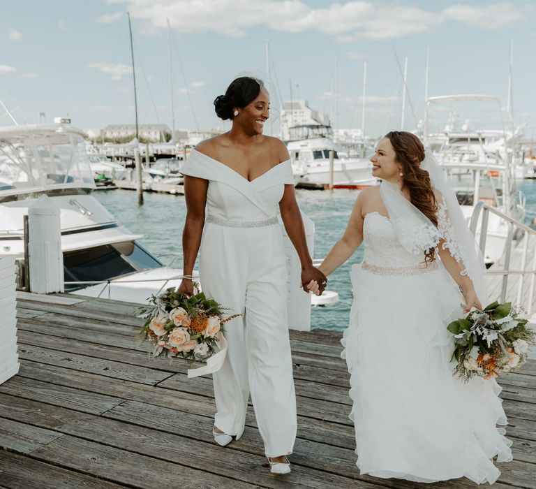 Black bride in an off the shoulder jumpsuit and bride in a strapless wedding dress with full skirt holding hands on the marina dock with boats in the background 