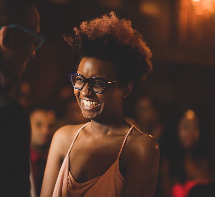 A bride in glasses laughs. She wears a blush pink dress and her hair os cropped to the sides but long and curly on top.