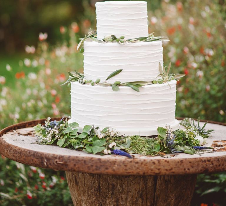 Three tier white buttercream iced cake on rustic table outdoors decorated with olive foliage