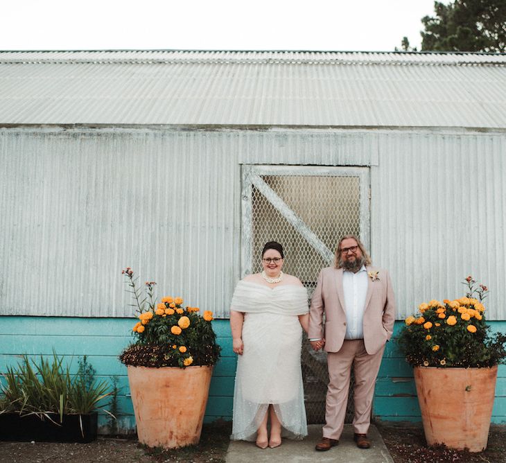 Portrait of the bride in a Bardot wedding dress with layer pearl tulle and groom in a matching pink trousers and jacket 