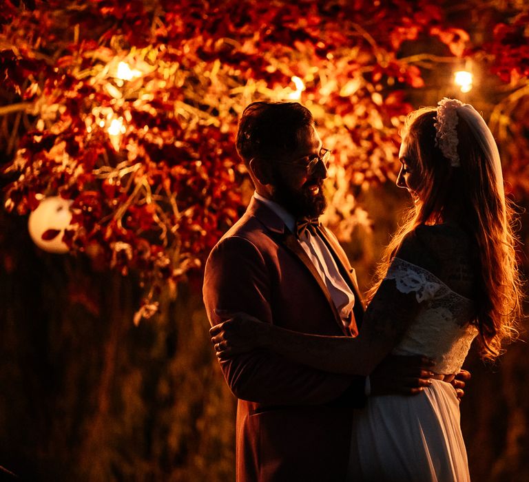 Bride & groom stand under trees lit by fairy lights