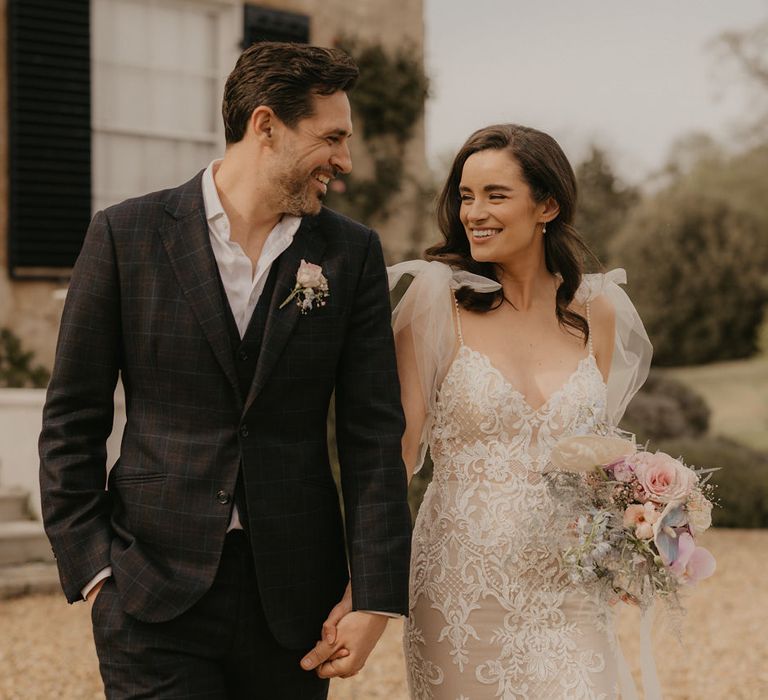 Bride in a fitted lace wedding dress with tulle shoulder detail holding a pastel wedding bouquet smiling at her groom in a check suit 