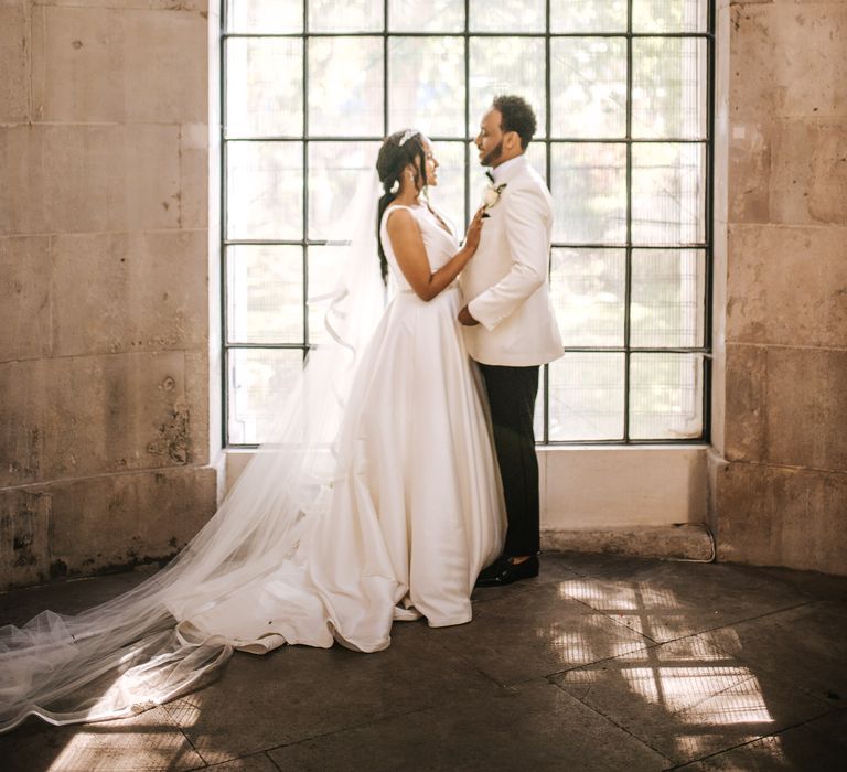 Bride in a princess wedding dress with full skirt and groom in a white tuxedo jacket standing in front of a window 