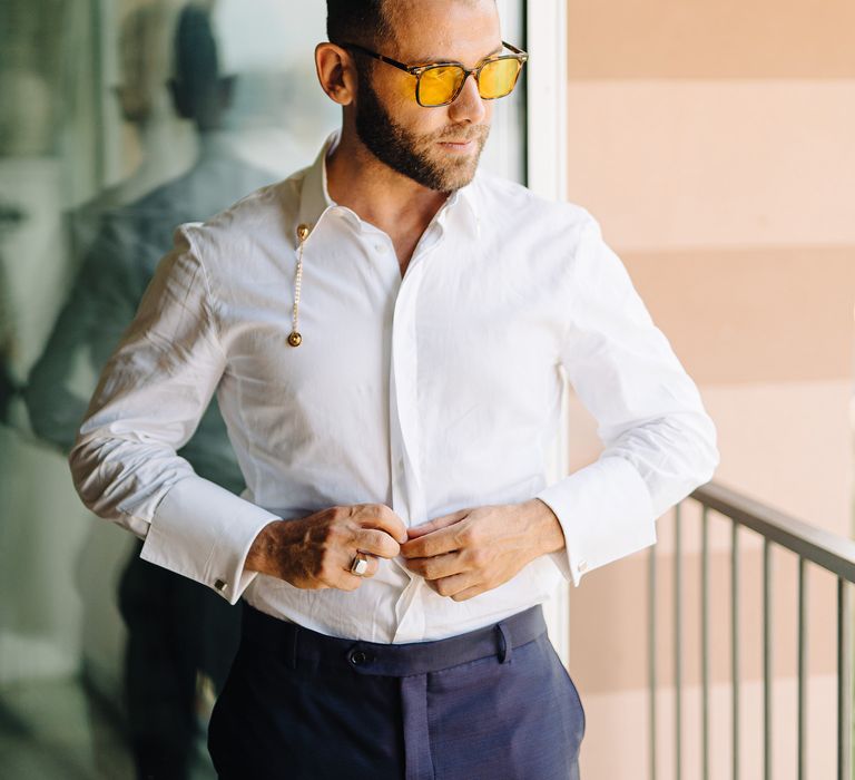 Groom does up his shirt on the morning of his wedding whilst stood on balcony