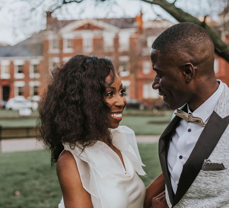 Smiling bride with curled hair in white Bec + Bridge satin wedding dress with shoulder bows smiles at groom in grey suit jacket with black lapels outside Bridge Community Church wedding