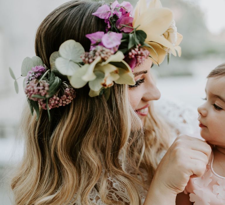 Bride with curled blonde hair and multicoloured flower crown touches chin of baby in pink dress with floral detailing at enchanted forest wedding in Italy