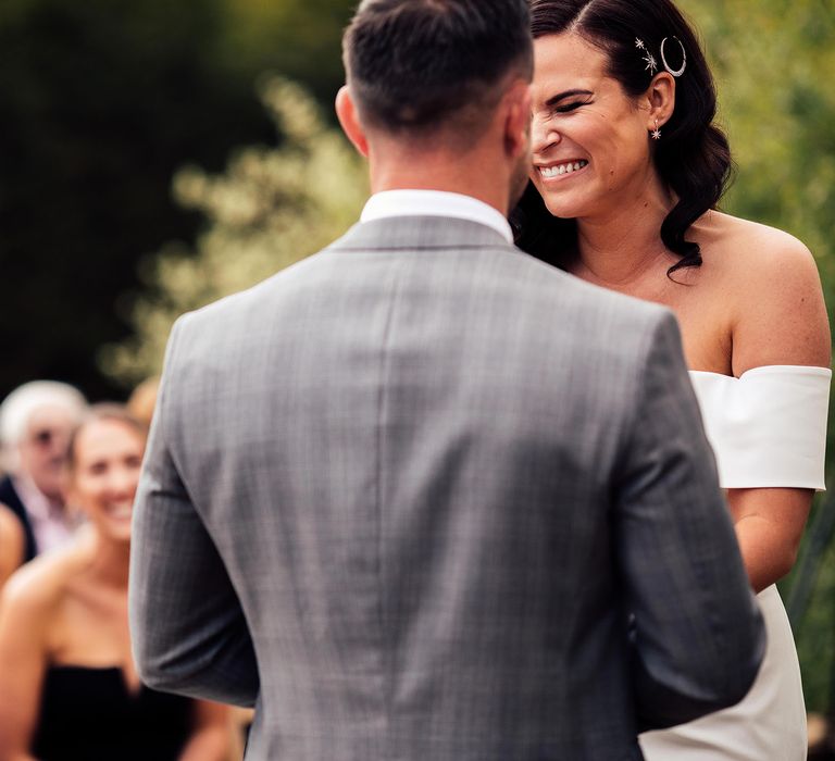 Bride in strapless Vagabond wedding dress with celestial hair clips laughs at the altar with groom in grey Hugo Boss suit 