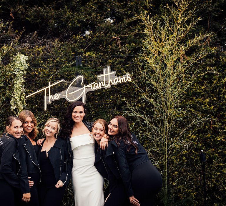 Bride in white strapless Vagabond dress and leather jacket stands under neon sign with maids of honours and lushers in personalised suede jackets