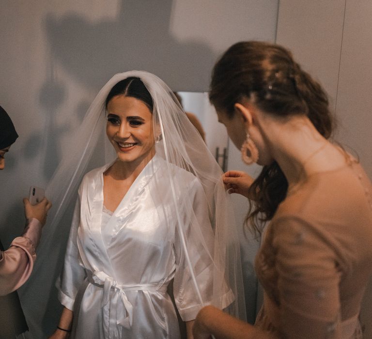 Bride has her veil applied by wedding party whilst wearing white silk dressing gown