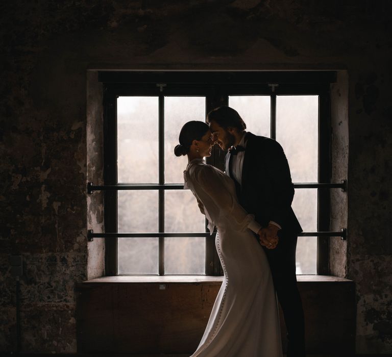 Bride & groom stand in front of window as the groom holds her hand and leans back