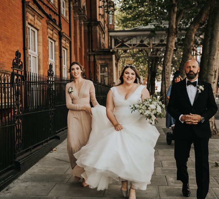 Bride walks through the streets of London as bridesmaid in blush pink dress with long sleeves holds her wedding gown