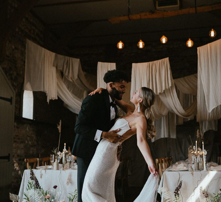 Groom in a tuxedo dancing with his bride in a sparkly wedding dress under drapes and festoon light ceiling decor 