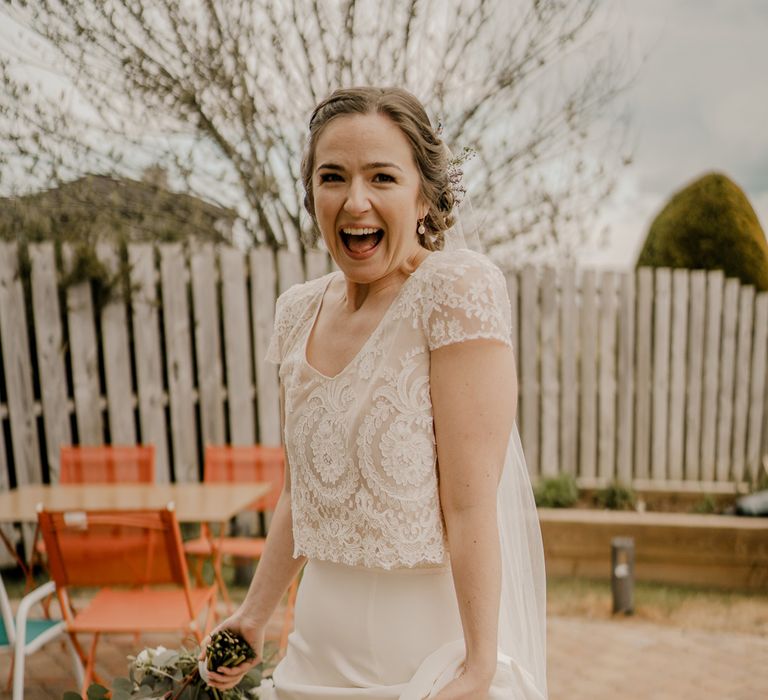 Smiling bride in lace top wedding dress with capped sleeves holds her satin skirt and bouquet before Dunluce Castle wedding 