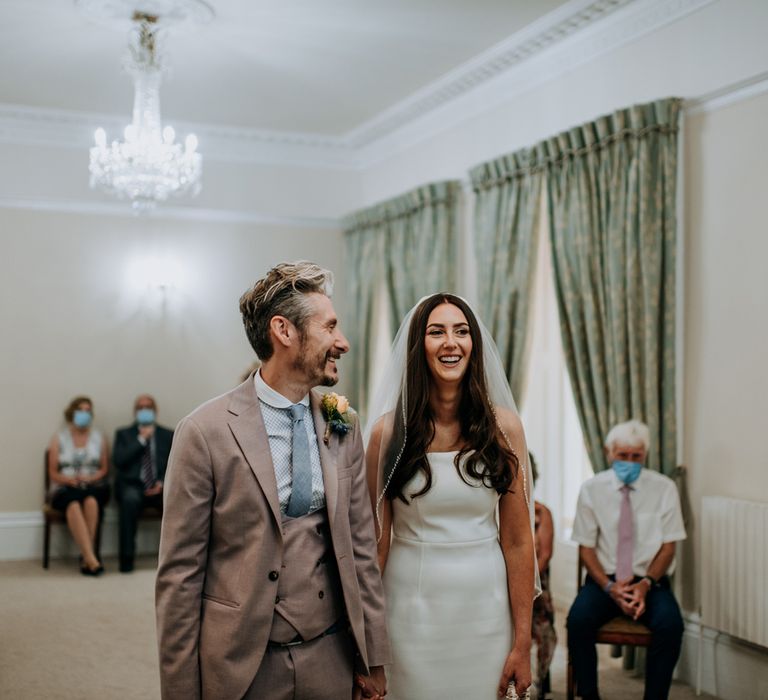 Bride in white strapless Rebecca Vallance Dress, veil and blue slingback heels carries ivory bridal bag whilst holding hands with groom in brown three piece Moss Bros suit and blue tie as they walk down the aisle at Harrogate wedding