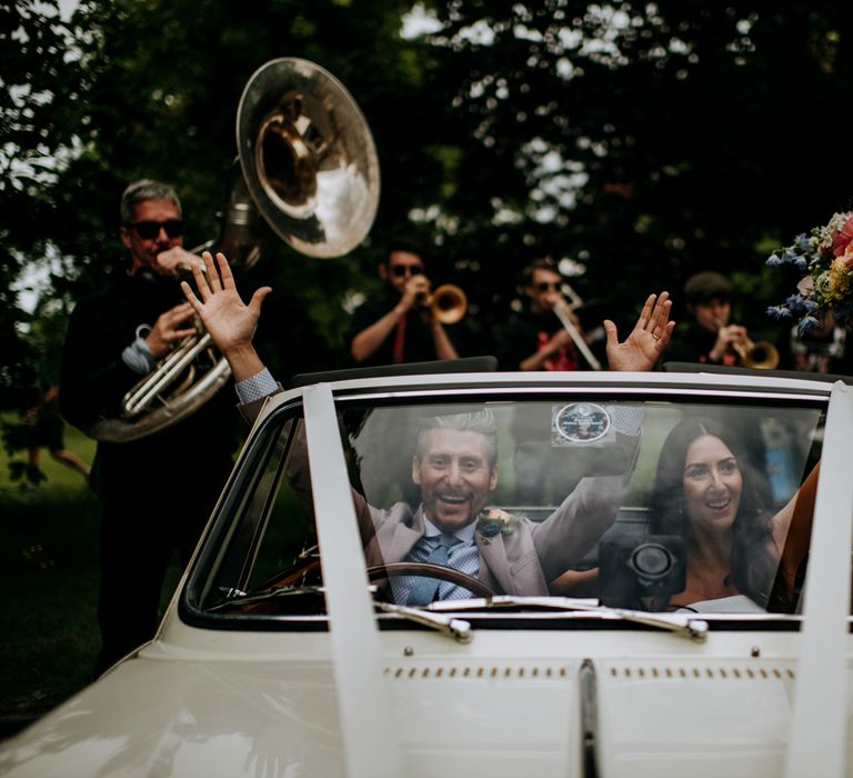 Bride and groom in vintage Triumph convertible surrounded by the New York Brass Band after Harrogate wedding