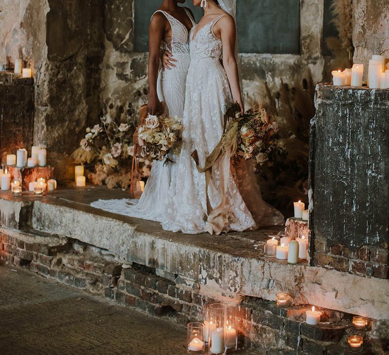 Two brides in appliqué and embellished wedding dresses standing at The Asylum altar surrounded by pillar candles and dried flower arrangements 