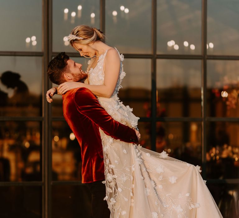 Groom in a red velvet tuxedo jacket lifting his bride up in a flower appliqué wedding dress and pearl headband in front of the Crittall windows at Botley Hill Barn wedding venue in Surrey 