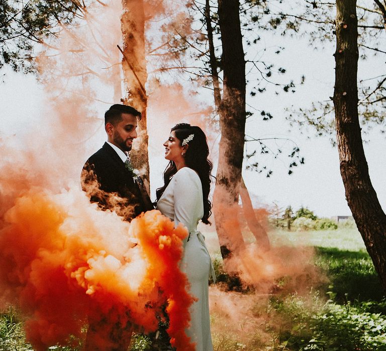 Orange smoke bomb wedding portrait with bride in a long sleeve wedding dress and groom in a navy three-piece suit 