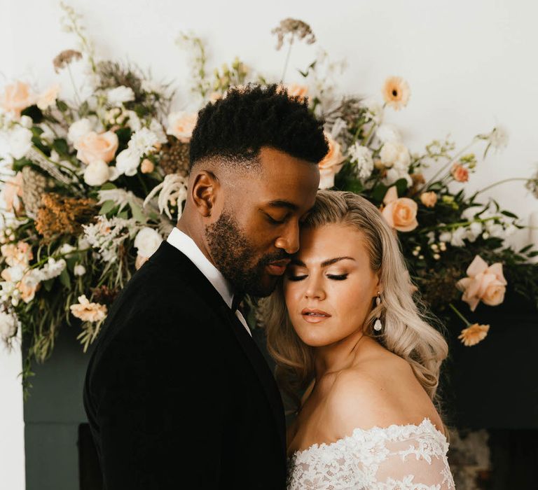 Bride and groom in front of a mantlepiece of white and peach florals, the bride is holding a bouquet of the same flowers, wearing an off the shoulder lace wedding dress. The groom is wearing a velvet suit.