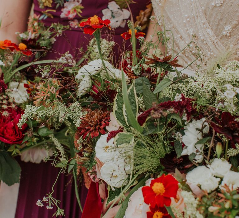 Bride in pearl wedding dress holds white and green wedding bouquets with red roses and foliage for barn wedding ceremony 