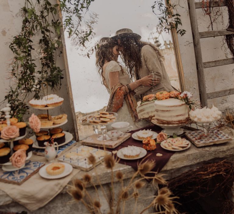 Bride & groom kiss in the reflection of rustic mirror as dessert buffet sits upon stone top