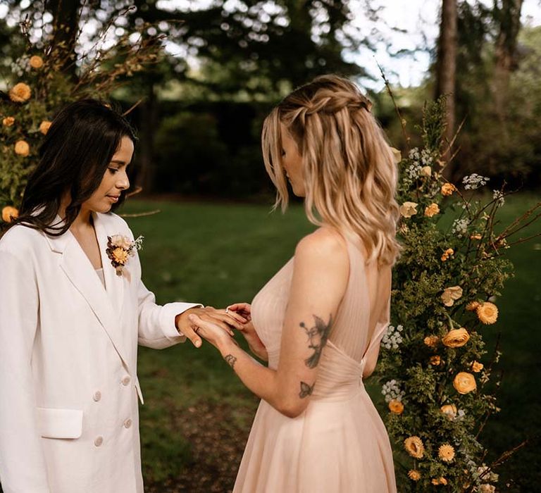 Bride in a pink tulle wedding dress putting a ring on her brides wedding finger at their outdoor wedding ceremony 