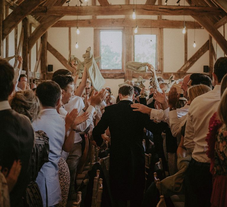 Groom in morning coat walks into high ceilinged beamed room with naked hanging bulbs as wedding guests applaud and wave napkins for wedding reception at Loseley Park in Surrey