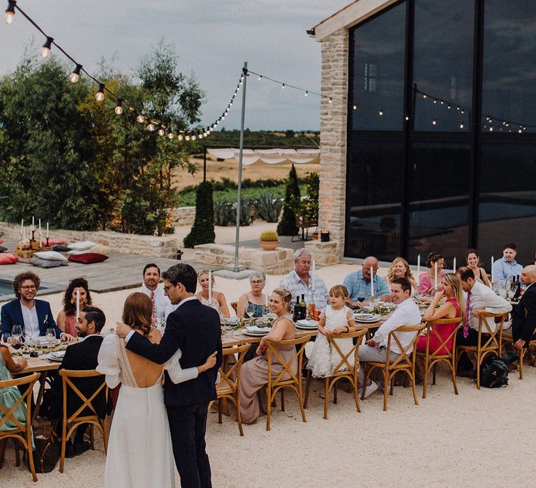 Wedding party sit outdoors at rustic table setting with bride & groom standing in front of them