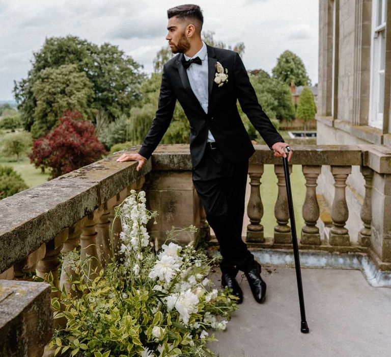 Groom in a black tuxedo with his walking stick standing on the balcony at Bourton Hall wedding venue 