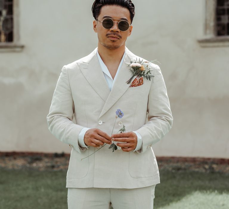 Groom in round sunglasses, shawl collared shirt and double-breasted linen suit stands outside church before Wasing Park wedding