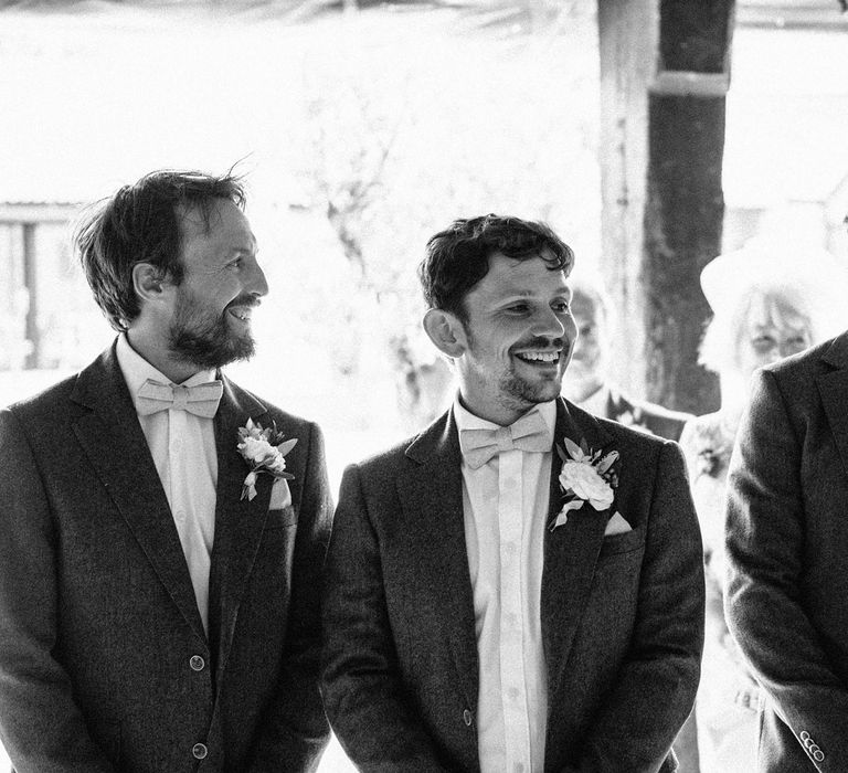 Groom and groomsmen in suit jackets, white shirts, bow ties and floral buttonholes wait at the alter for bride at Isle of Wight wedding