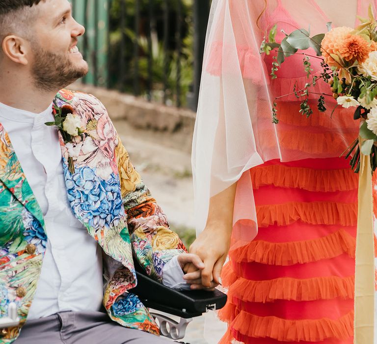 Bride in a coral wedding dress, cape veil and flower crown holding hands and smiling at her groom in a patterned blazer 