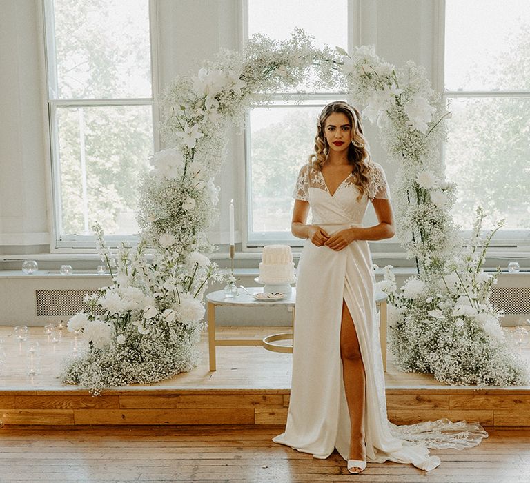 Bride with long wavy hair and red lipstick in a wrap wedding dress with lace detail standing in front of a white flower arch