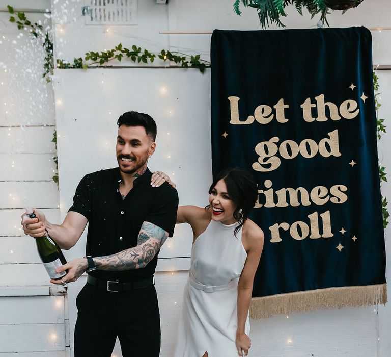 Groom in a black shirt and bride in a halterneck wedding dress with red lipstick popping champagne in front of a 'let the good times roll' banner 