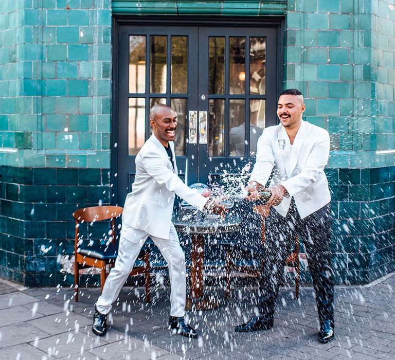 Two groom in a suite suit and black leather trousers popping champagne 