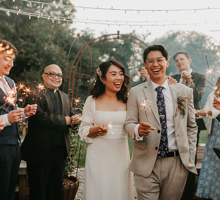 Bride & groom hold sparklers as they walk through sparkler exit surrounded by wedding guests outdoors