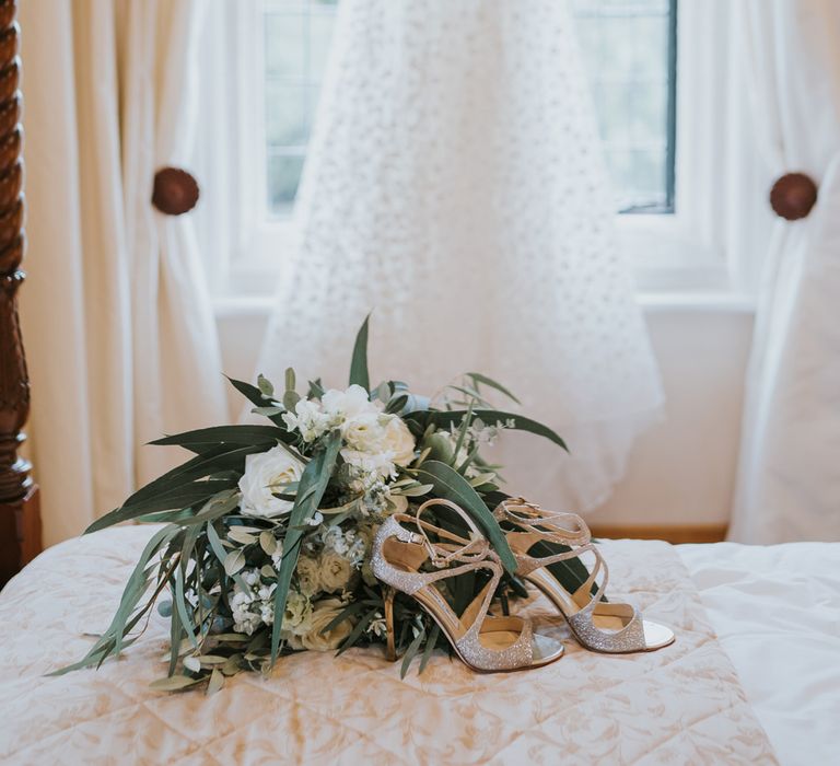 Cami strap white Pronovias wedding dress hung on window behind Jimmy Choo wedding shoes and green and white rose bridal bouquet