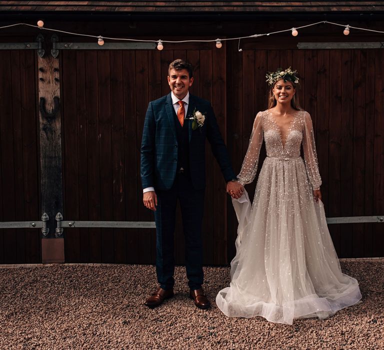 Smiling bride in a line mesh long sleeve Julie Vino wedding dress and flower crown stands holding hands with groom in blue checked three piece suit, orange tie and floral buttonhole