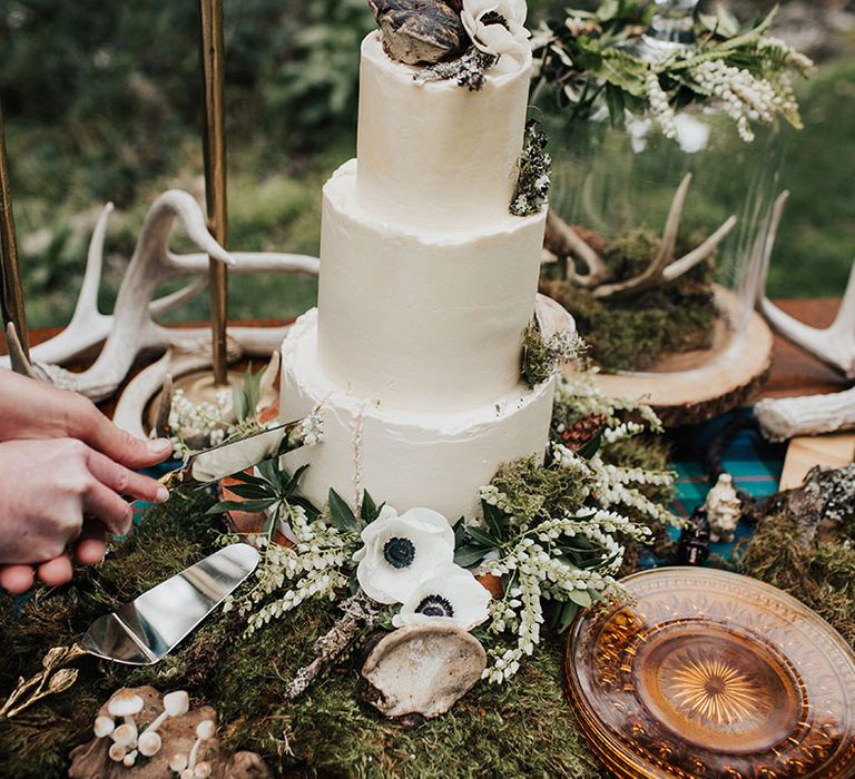 Bride and groom cutting the buttercream wedding cake surrounded by moss and anemone decor 