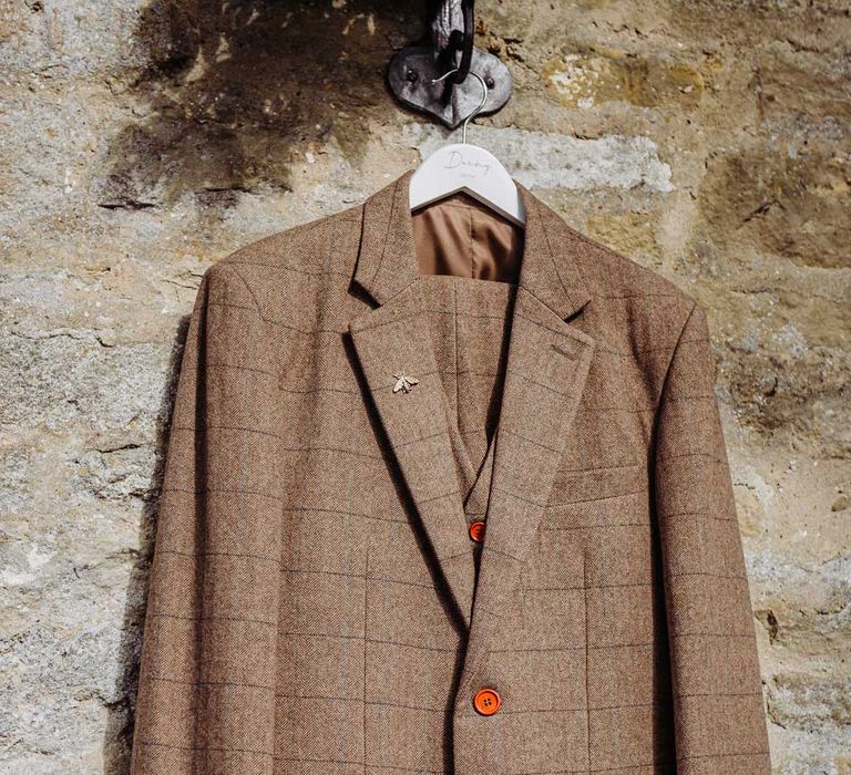 Brown tweed grooms suit with orange buttons hanging on stone wall