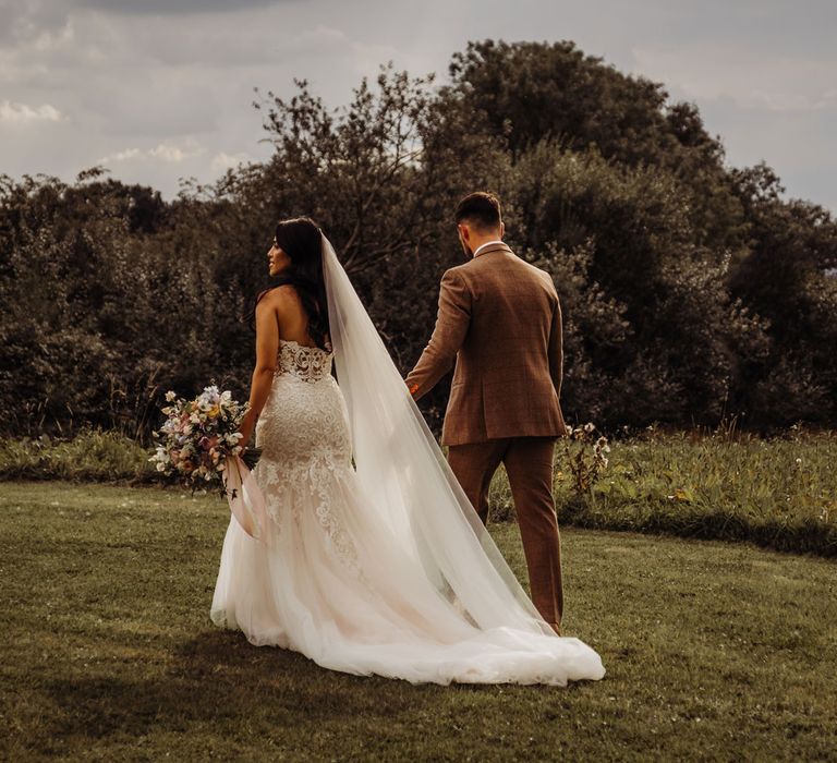 Bride in lace and tulle wedding dress with train and veil holding multicoloured bridal bouquet walks through field with groom in brown tweed suit for Cotswolds wedding