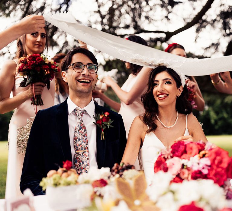 Bride in v neck wedding dress wearing feathered headband and pearl necklace sits with groom in dark suit, floral Gucci tie and floral buttonhole during traditional sugar ceremony