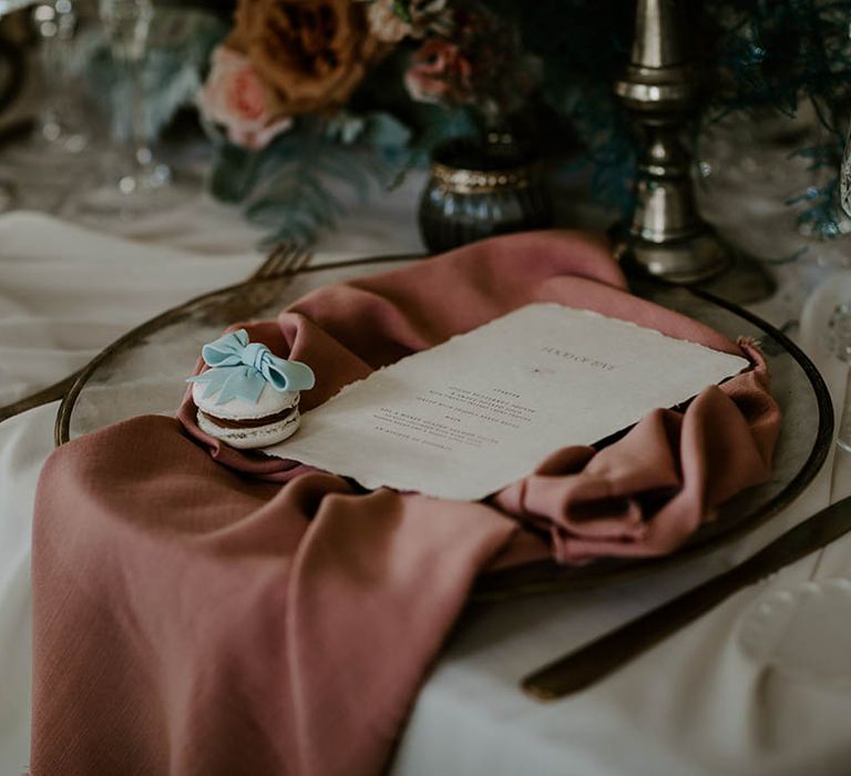 Place setting with gold rimmed glass charger plate, pink napkin, and macaroon wedding favour