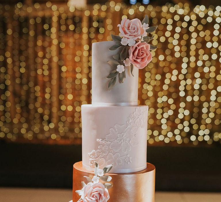 Four-tier wedding cake with white frosting and rose gold/copper frosting with decorative roses and details