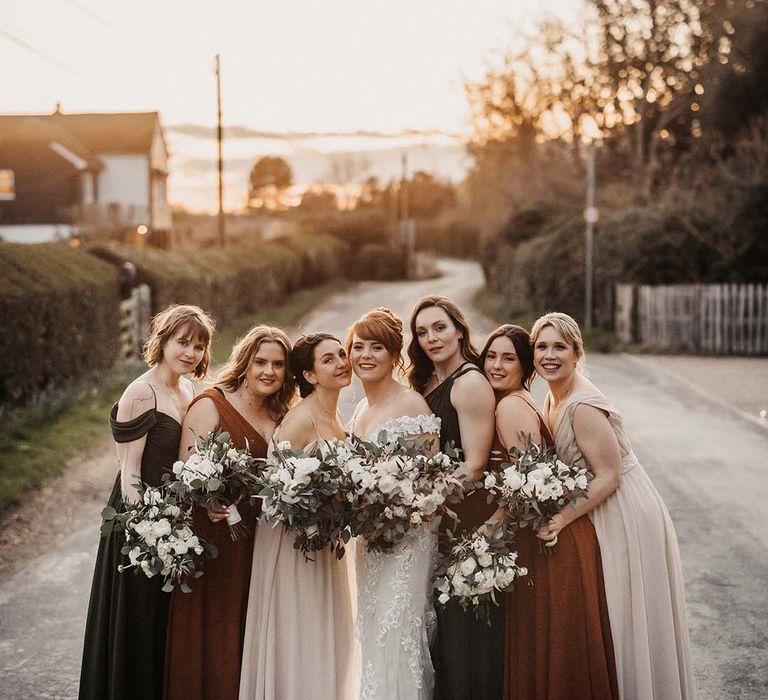 Bridesmaids in brown, rust and cream coloured dresses with various necklines stand with bride in lace wedding dress