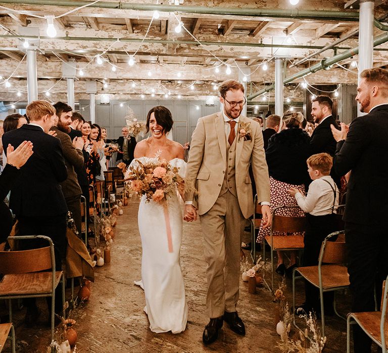 Groom in beige three piece suit and bride in Alexandra Grecco gown walk back down the aisle as a married couple 