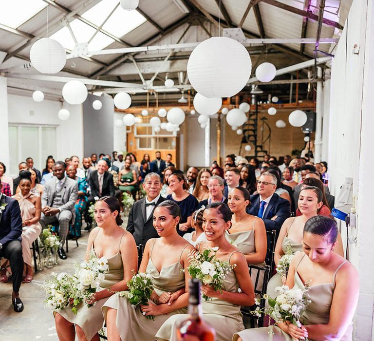 Bridesmaids with sleek hair and mint green bridesmaid dresses hold white flower bouquets at industrial wedding venue with groomsmen and other guests 