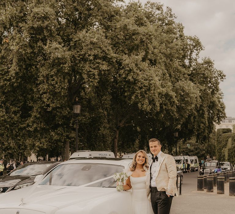 Groom in patterned custom gold tuxedo with bride in ruffle sleeve wedding dress stands next to their white Rolls Royce wedding car 