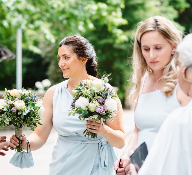 Bridesmaids in pale baby blue dresses with matching face masks holding white and purple bouquets 