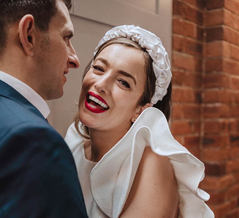 Bride wearing bright red lipstick smiles brightly wearing white ruffle headband and Jesus Peiro wedding dress held by groom in blue suit 